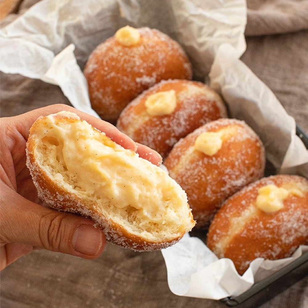 Hand holding a Rhukra Cake House banana bombolone doughnut with creamy filling and sugar coating in a paper-lined box.