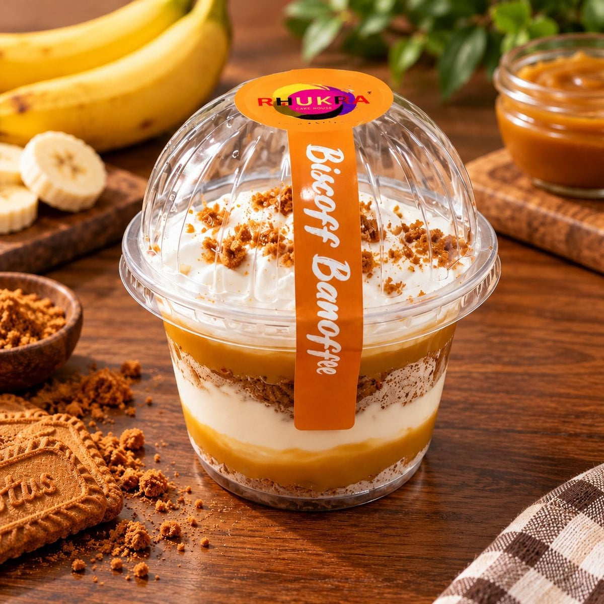 Rhukra Cake House Biscoff Banoffee dessert in a clear plastic cup with layers of cream, caramel, and cookie crumbs.
