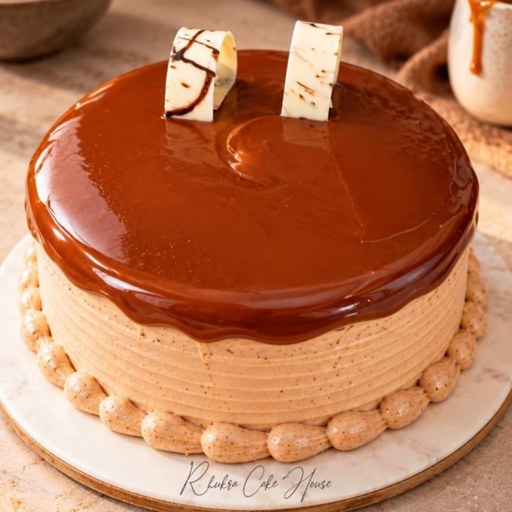 Rhukra Cake House Biscoff Caramel cake with a smooth caramel glaze, piped frosting border, and white chocolate curls.