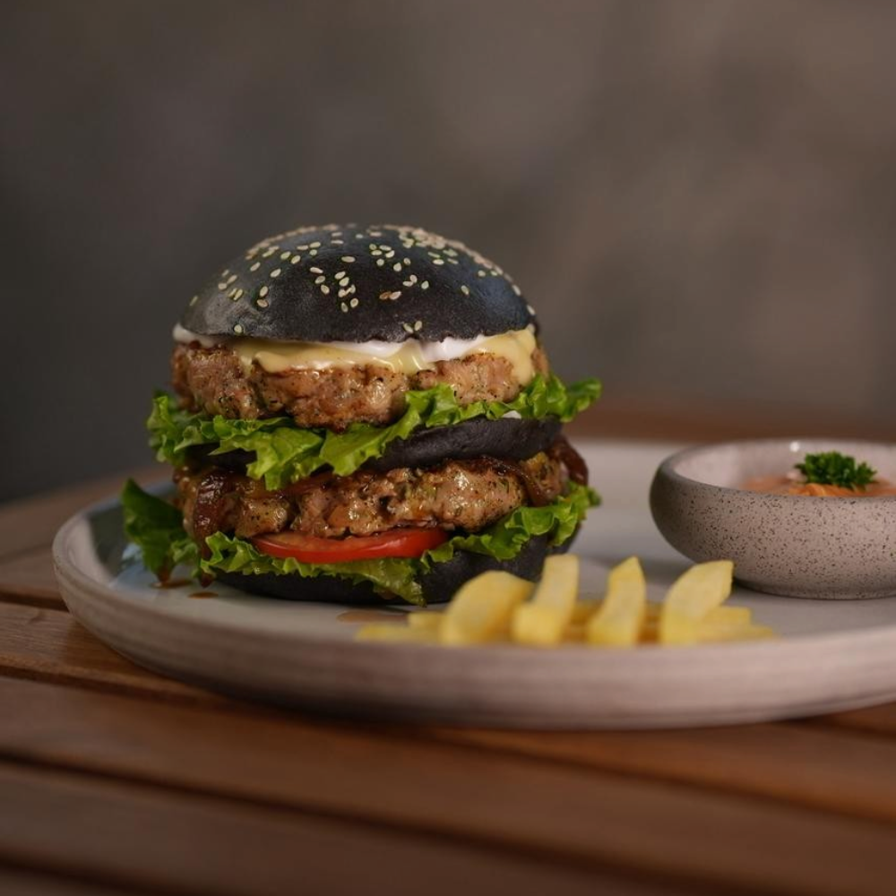 Rhukra Cake House double chicken patty burger with black sesame bun, lettuce, tomato, and cheese served with fries.