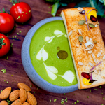 Rhukra Cake House cream of broccoli soup in a grey bowl with toasted bread, fresh broccoli, tomatoes, and almonds.