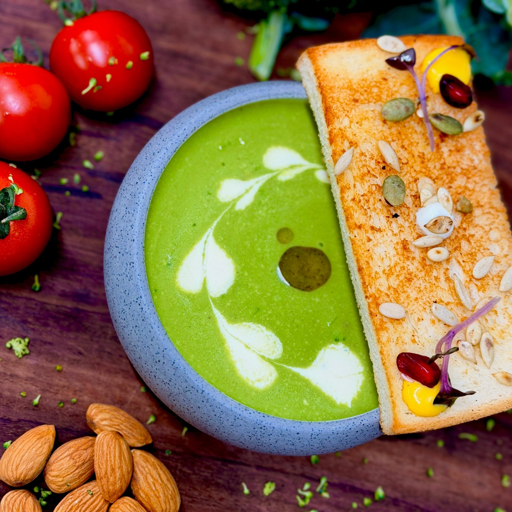 Rhukra Cake House cream of broccoli soup in a grey bowl with toasted bread, fresh broccoli, tomatoes, and almonds.