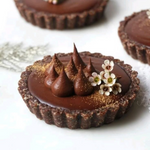 Rhukra Cake House chocolate tart with ganache swirls, edible gold dust, and small white flowers on a white surface.